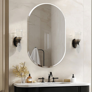 An oval wall mirror with a thin silver frame hangs above a white vanity with a black faucet. The bathroom wall is light beige marble, and two matching wall sconces flank the mirror.