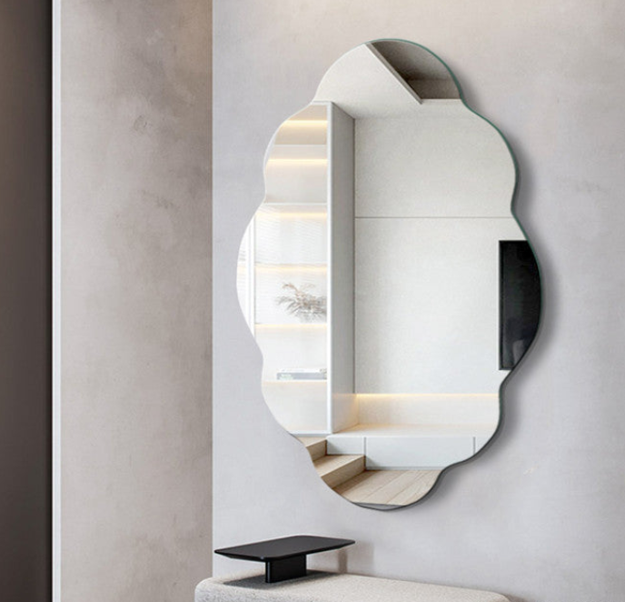 An organic-shaped wall mirror with a scalloped edge, mounted on a light gray textured wall above a small black side table. The mirror reflects a modern interior with built-in shelves and warm lighting.