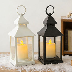 Two decorative lanterns with lit candles, one white and one black, placed on a surface covered with white faux snow in front of a neutral indoor background.