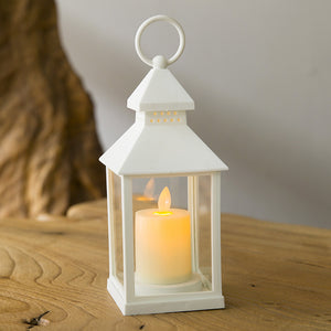 A white lantern-style candle holder with a lit cream pillar candle inside, placed on a wooden surface against a softly blurred neutral background.