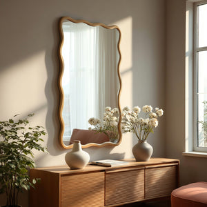 A large rectangular wall mirror with a wavy wooden frame hangs on a beige wall above a wooden console table. The scene is styled with two ceramic vases containing white flowers, a potted plant, and natural sunlight streaming in from a window.