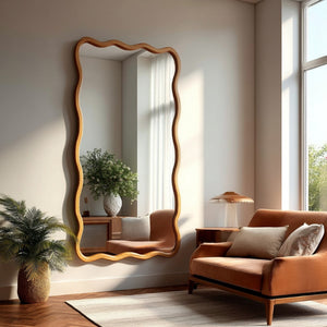 A tall, wavy-edged wooden wall mirror with a natural oak frame, styled in a warm, modern living room next to a rust-orange armchair and a potted palm.