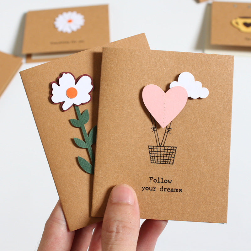 A hand holds two small kraft paper cards with layered paper cutouts. The front card shows a pink heart hot air balloon, white clouds, and the text 'Follow your dreams.