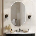An oval wall mirror with a thin silver frame hangs above a white vanity with a black faucet. The bathroom wall is light beige marble, and two matching wall sconces flank the mirror.