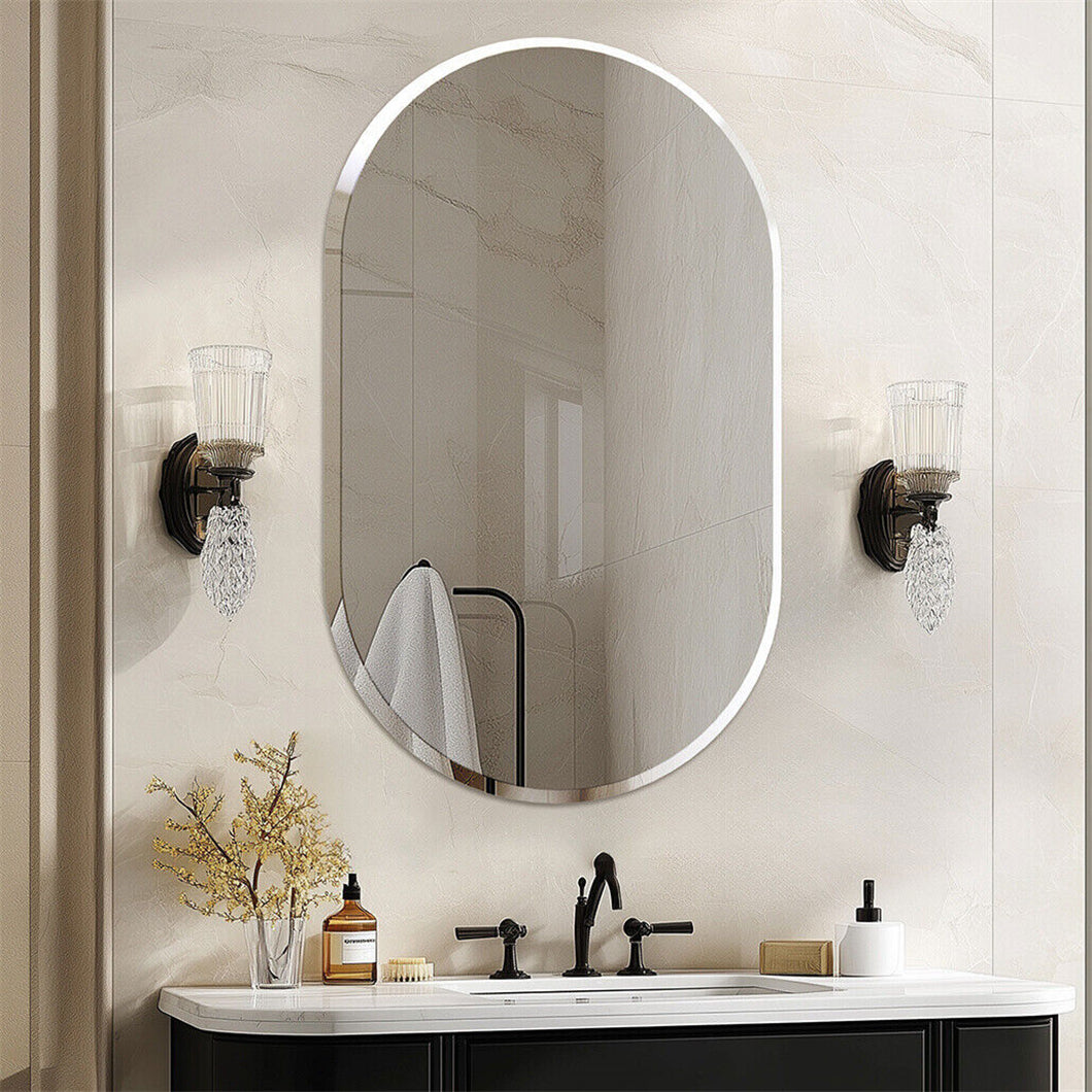 An oval wall mirror with a thin silver frame hangs above a white vanity with a black faucet. The bathroom wall is light beige marble, and two matching wall sconces flank the mirror.