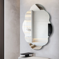 An organic-shaped wall mirror with a scalloped edge, mounted on a light gray textured wall above a small black side table. The mirror reflects a modern interior with built-in shelves and warm lighting.