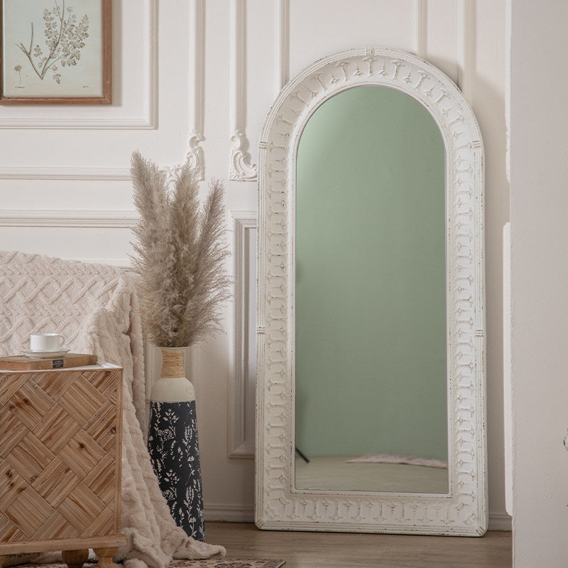 An arched full-length floor mirror with an ornate distressed white frame, leaning against a white paneled wall in a styled room next to a woven side table and a vase of pampas grass.
