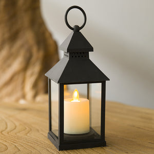 A black metal lantern with a glass front and a hanging loop on top, holding a lit white candle inside. The lantern sits on a wooden surface with a softly blurred background.