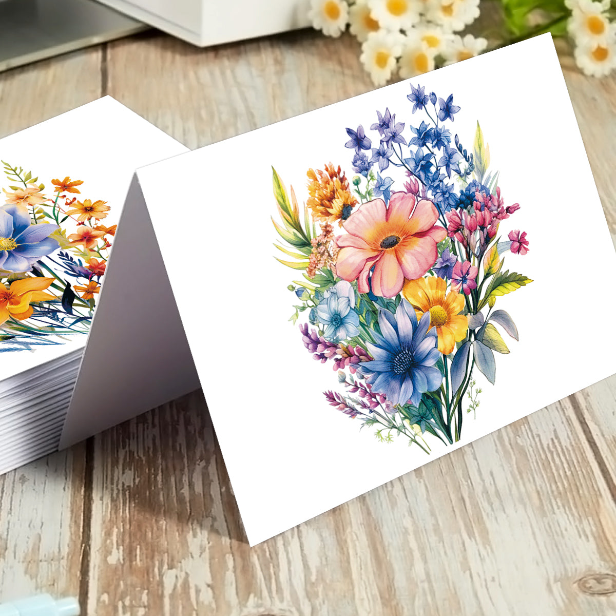 A white greeting card standing upright on a rustic wooden surface, printed with a colorful watercolor-style bouquet of flowers. A stack of similar cards sits to the left, and blurred flowers appear in the background.
