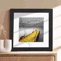 A framed art print featuring a yellow wooden dock extending into a calm lake with snow-capped mountains in the background, displayed on a wooden surface next to a white ceramic vase.
