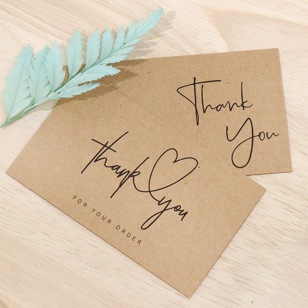 Two brown kraft paper thank you cards with black script text, displayed on a light wood surface with a decorative mint green leaf.